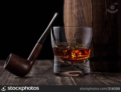Glass of whiskey with ice cubes and vintage smoking pipe next to wooden barrel. Cognac and brandy drink