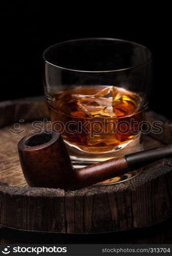 Glass of whiskey with ice cubes and vintage smoking pipe on top of wooden barrel. Cognac and brandy drink