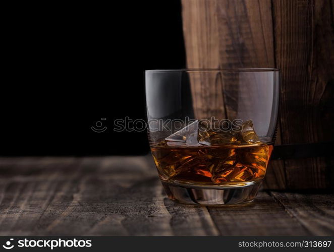 Glass of whiskey with ice cubes next to wooden barrel. Cognac and brandy drink