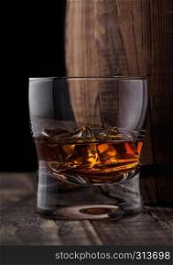 Glass of whiskey with ice cubes next to wooden barrel. Cognac and brandy drink