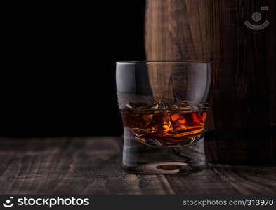 Glass of whiskey with ice cubes next to wooden barrel. Cognac and brandy drink