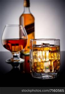 Glass of whiskey with ice on a dark background. In the background a glass with whiskey and a bottle of whiskey. Closeup of a whiskey boil with ice. Background full bottle of whiskey and a filled glass