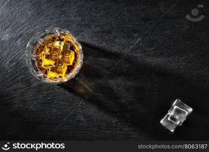 Glass of whiskey with ice on black textural background shot from above. Glass of whiskey with ice on black textural background