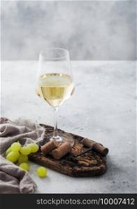Glass of white homemade wine with corks, corkscrew and grapes on wooden board with linen cloth on light table background.