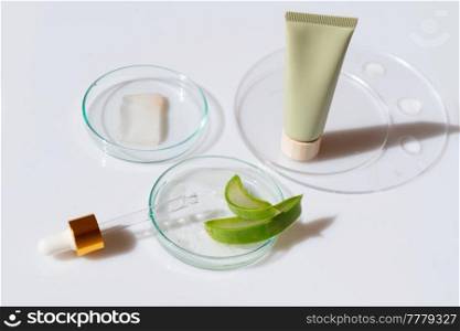 Glass petri dish with transparent pure serum for skin care with cosmetic tube and aloe vera pieces on white background, top view. Concept laboratory tests and research, making and testing cosmetic. Glass petri dish with transparent pure serum for skin care