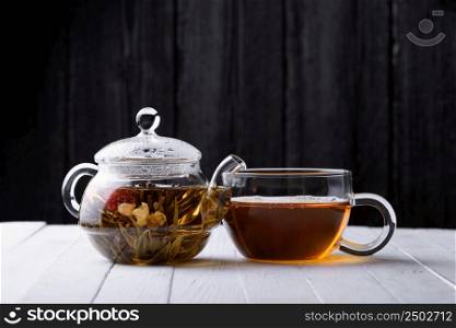 Glass teapot with blooming green tea and cup of tea on white wooden table and dark background still life