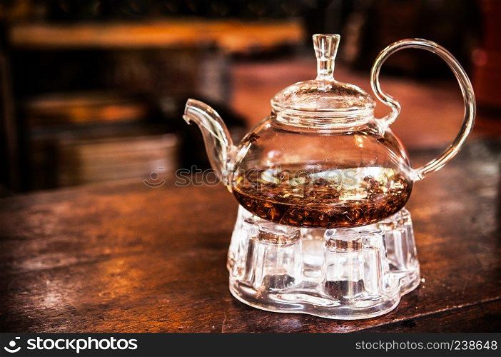 Glass teapot with tea leaves on wooden table and dark background in warm light