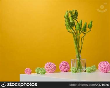 glass transparent vase with green dried flowers on a white table, yellow background