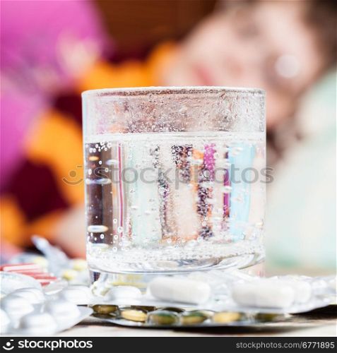 glass with dissolved drug in and pills on table close up and sick girl with scarf around her neck on sofa in living room on background
