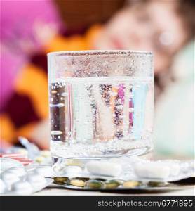 glass with dissolved drug in and pills on table close up and sick girl with scarf around her neck on sofa in living room on background