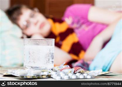 glass with dissolved drug in water and pile of pills on table close up and sick girl with scarf around her neck on sofa in living room on background
