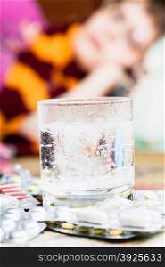 glass with dissolved medicament in water and pile of pills on table close up and sick girl with scarf around her neck on sofa in living room on background