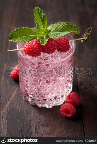 Glass with ice of cold summer pink lemonade cocktail with raspberries, mint and raw berries on wooden background. Macro