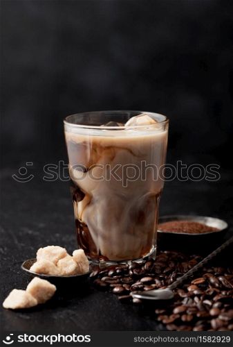 Glass with iced coffee with beans and cane sugar with ground coffee and long spoon on black table background.