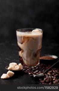 Glass with iced coffee with beans and cane sugar with ground coffee and long spoon on black table background.