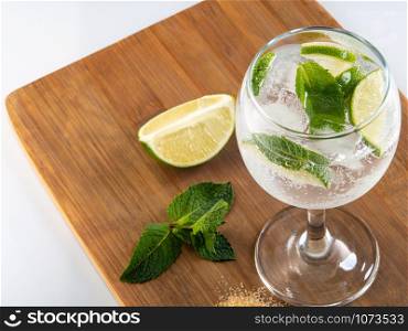 Glass with refreshing lemonade, lime slices and mint on a kitchen cutting board. Cooked mojito on a wooden tray and cocktail ingredients. Top view.