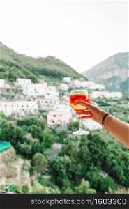 Glass with Spritz Aperol alcohol drink background of beautiful old italian village on Amalfi coast. Female hand holding glass with Spritz Aperol alcohol drink background of beautiful old italian village on Amalfi coast