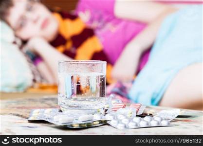 glass with water and pills close up and sick girl with scarf around her neck on sofa in living room on background