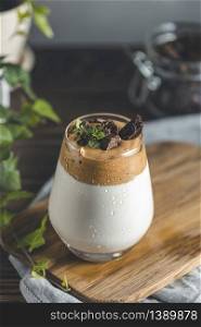 Glass with water drops of Iced Dalgona Coffee, a trendy fluffy creamy whipped coffee, decorated by thyme and dark chocolate. Dark rustic style.