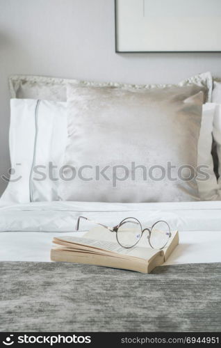 Glassed and book on bed with velvet finished pillows on background