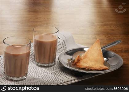 Glasses of chai on newspaper with plate of samosa over a wooden surface