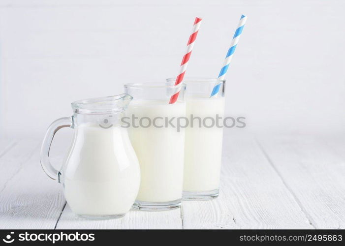 Glasses of milk with stripped blue and red paper straws and jug of milk on white wooden table still life