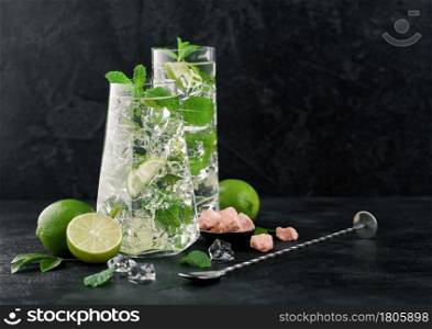 Glasses of Mojito cocktail with ice cubes mint and lime on black board with spoon and fresh limes with cane sugar. Best summer cocktail.