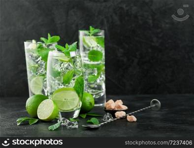 Glasses of Mojito refreshing summer cocktail with ice cubes mint and lime on black board with spoon and fresh limes with cane sugar. Sparkling refreshing water. Space for text
