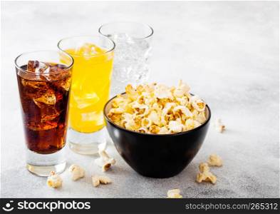 Glasses of soda drink with ice cubes and black bowl of popcorn snack on stone kitchen background.
