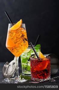 Glasses of spritz,mojito and negroni cocktails with ice cubes and lime and orange slices with mint leaf and black straw on dark background with strainer and spoon.