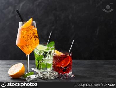 Glasses of spritz,mojito and negroni cocktails with ice cubes and lime and orange slices with mint leaf and black straw on dark background with strainer. Space for text