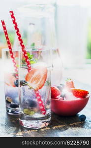 Glasses with berries lemonade with red straw and ice cubes on kitchen table over garden background. Home scene with summer drinks.