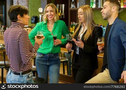 Gleeful diverse man and women with alcohol beverages smiling and talking while spending weekend day in bar together. Happy friends chatting in a beautiful bar