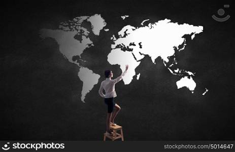 Global business interaction. Rear vew of businesswoman climbing chair and touching world map on wall
