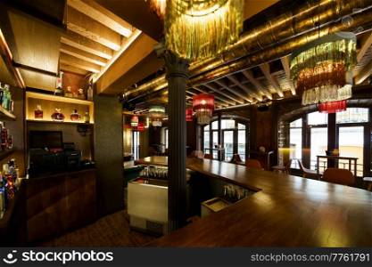 Glowing chandeliers illuminating spacious contemporary restaurant with brown wooden furniture and cozy atmosphere. Interior of modern restaurant with wooden furniture