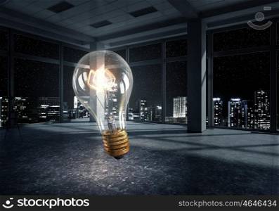 Glowing light bulb in modern office. Glass light bulb against room with large window looking on city