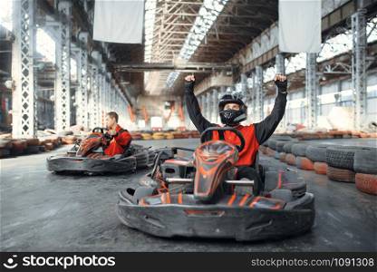 Go kart racer in helmet raised his hands up, winner, front view, karting auto sport indoor. Speed race on close go-kart track with tyre barrier. Fast vehicle competition, hot pursuit
