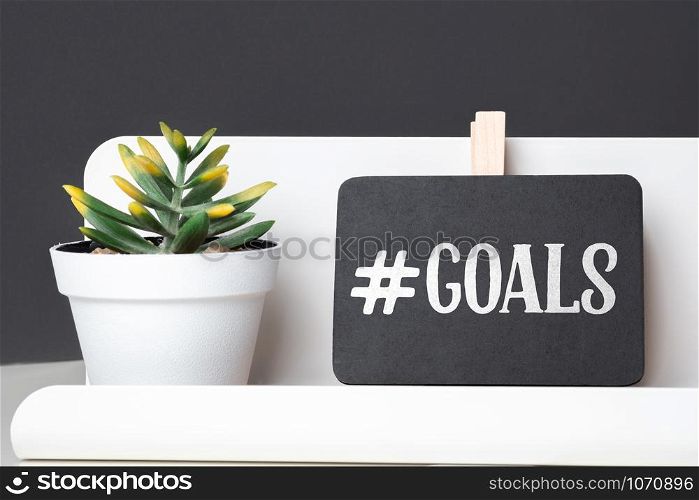 Goals on blackboard on pencil box and green plant in white pot on table and dark grey wall in office desk.business planning