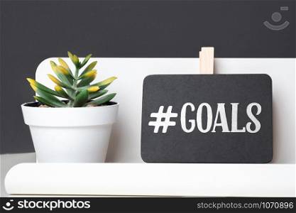 Goals on blackboard on pencil box and green plant in white pot on table and dark grey wall in office desk.business planning