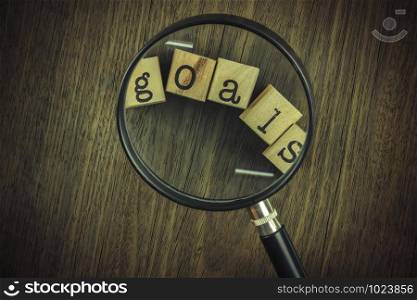 Goals setting strategy for business management. Magnifying glass focus on goals word typography over blocks and wooden table background, vintage and retro style.