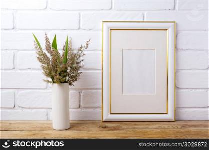 Gold decorated frame mockup with grass and green leaves in cyli. Gold decorated frame mockup with meadow grass and green leaves in cylinder vase near painted brick wall. Empty frame mock up for presentation artwork. Template framing for modern art.