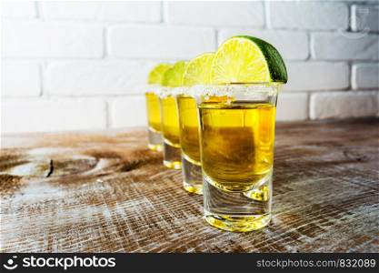 Gold Mexican tequila shots with lime slices on the rustic wooden background and painted brick wall.