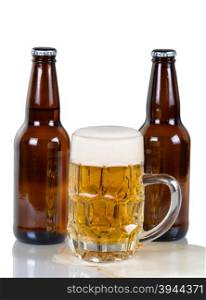 Golden colored beer flowing out of glass mug with two full bottles in background. Isolated on white with reflection.