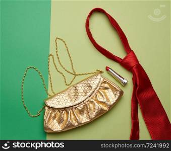 golden fashion handbag on a chain and a red silk tie on a green background, top view