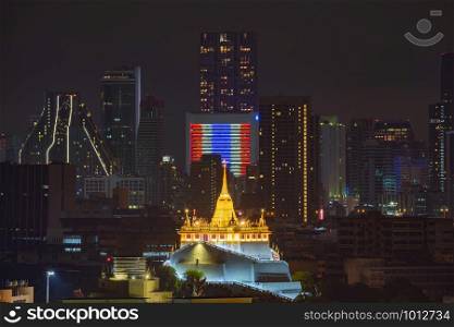 Golden Mount or Wat Saket, Phukhao Thong, and skyscraper buildings with Thailand flag at night in Bangkok City, Thailand. Buddhist temples.