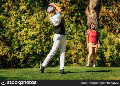 Golfing couple. Man teeing off with the driver, lady in the back, following the ball in flight