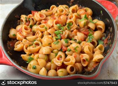 Gomiti elbow pasta shells in arrabbiata tomato, garlic and chili sauce, garnished with chopped parsley, cooking in a frying pan.