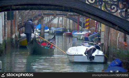 Gondola is sailing under the bridge in Venice, Italy. Multiple boats are parked at both sides of the water canal.