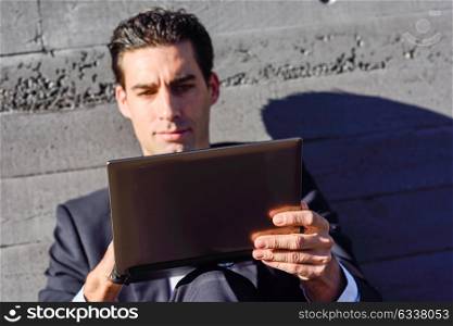Good looking businessman using a laptop computer sitting in the street. Man wearing blue suit and tie in urban background.