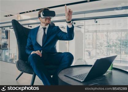 Good-looking CEO man in formal suit testing new product of company in virtual reality gaming technology, wearing VR headset goggles, gesturing with forefinger up in air, while sitting in office lobby. Well-groomed CEO man in formal suit wearing VR headset goggles, while sitting in office lobby